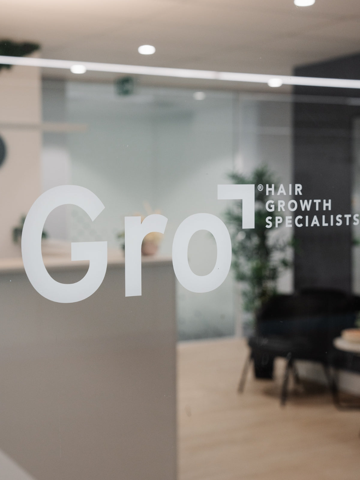 A modern office space with a glass door displaying the company's name, "Gro Hair Growth Specialists." The interior shows a few plants, chairs, and a well-lit, minimalist design, suggesting a professional and welcoming environment.