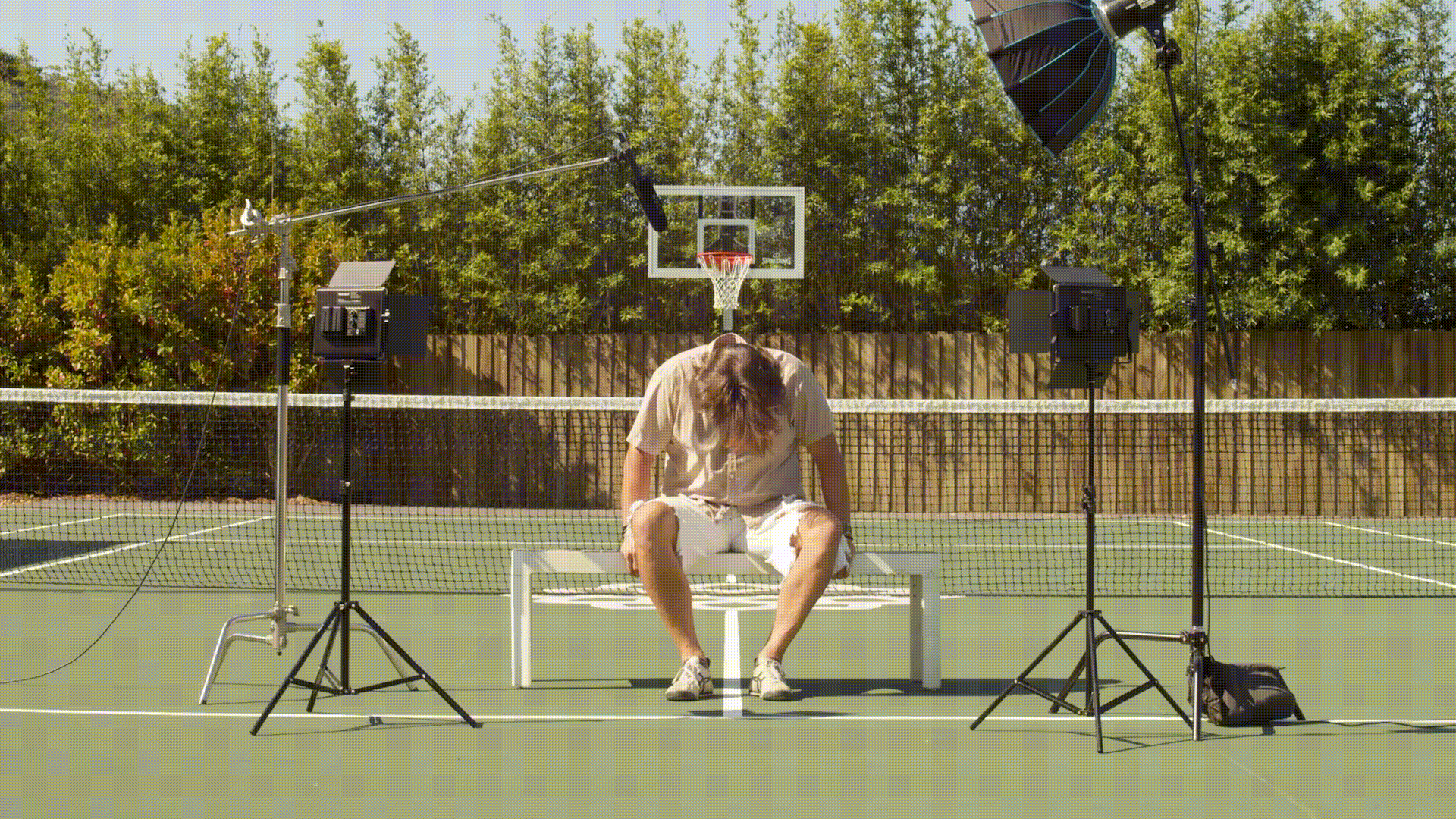 A man sitting on a bench with his head down on a tennis court. Surrounding him are professional studio lights and microphones pointed towards him, suggesting a photo or video shoot. A basketball hoop is visible in the background.