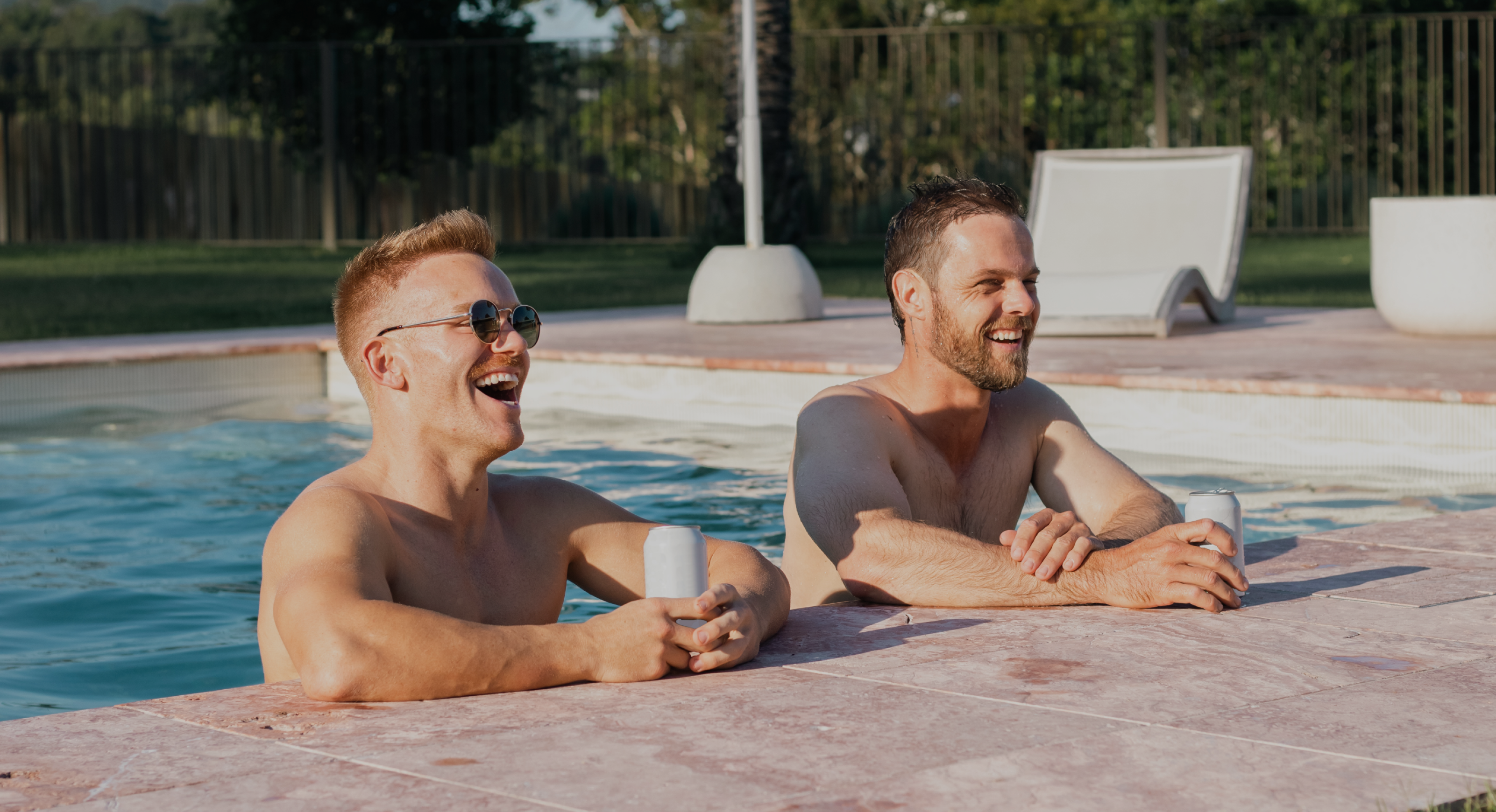 Two men are leaning on the edge of a swimming pool, smiling and holding cans. One of them, on the left, is wearing sunglasses. The background features greenery and an empty lounge chair. The sun is shining brightly, creating a relaxed and cheerful atmosphere.
