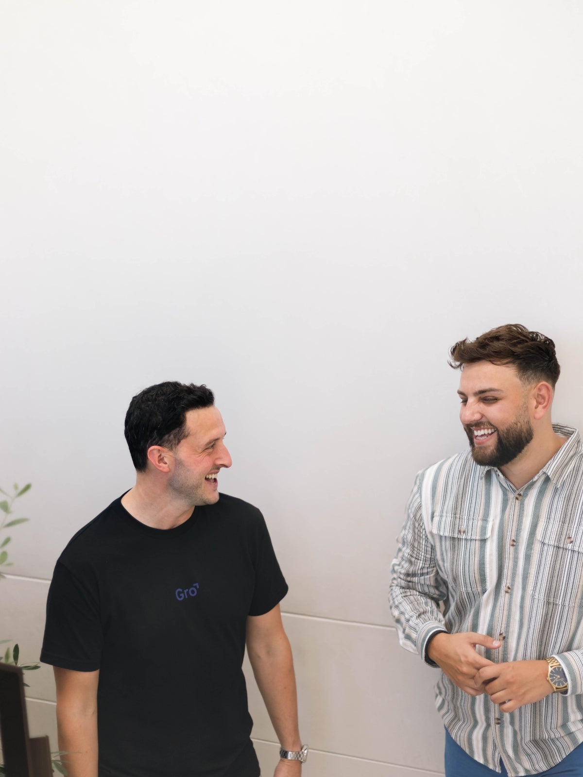 Two men are standing and laughing together against a plain white background. The man on the left is wearing a black T-shirt and the man on the right is wearing a striped button-up shirt. They appear to be in a relaxed and friendly conversation.