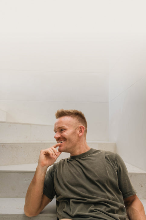 A man with short hair and a trimmed beard sits casually on a white staircase, wearing a grey t-shirt. He is leaning back with his left arm resting on a step, smiling and looking to his right, creating a relaxed and cheerful atmosphere.