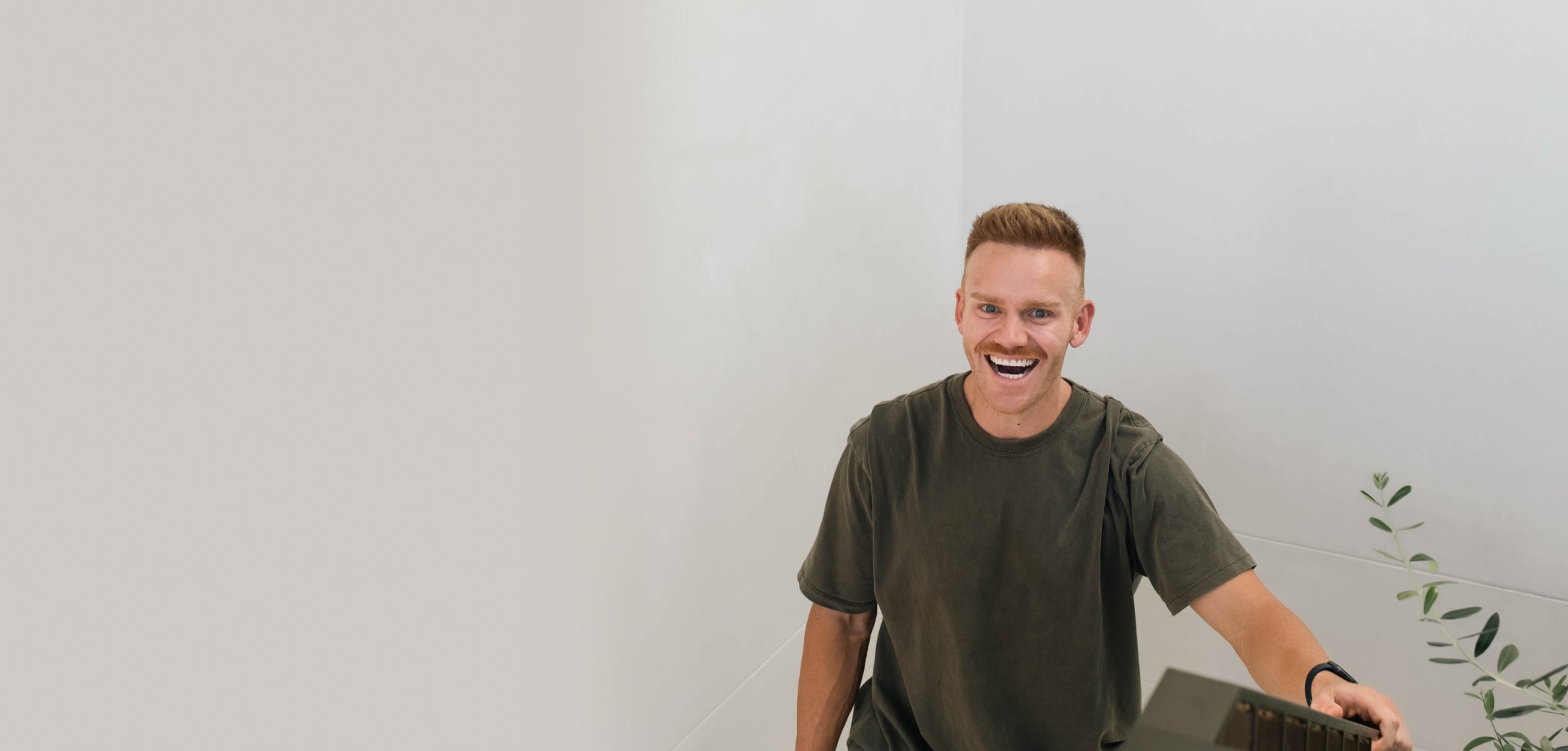 A person with short hair and a mustache sits at a desk or table, smiling warmly at the camera. They are wearing a green T-shirt and an armband, with a small plant visible on the right side of the image. The background is clean and minimalistic.