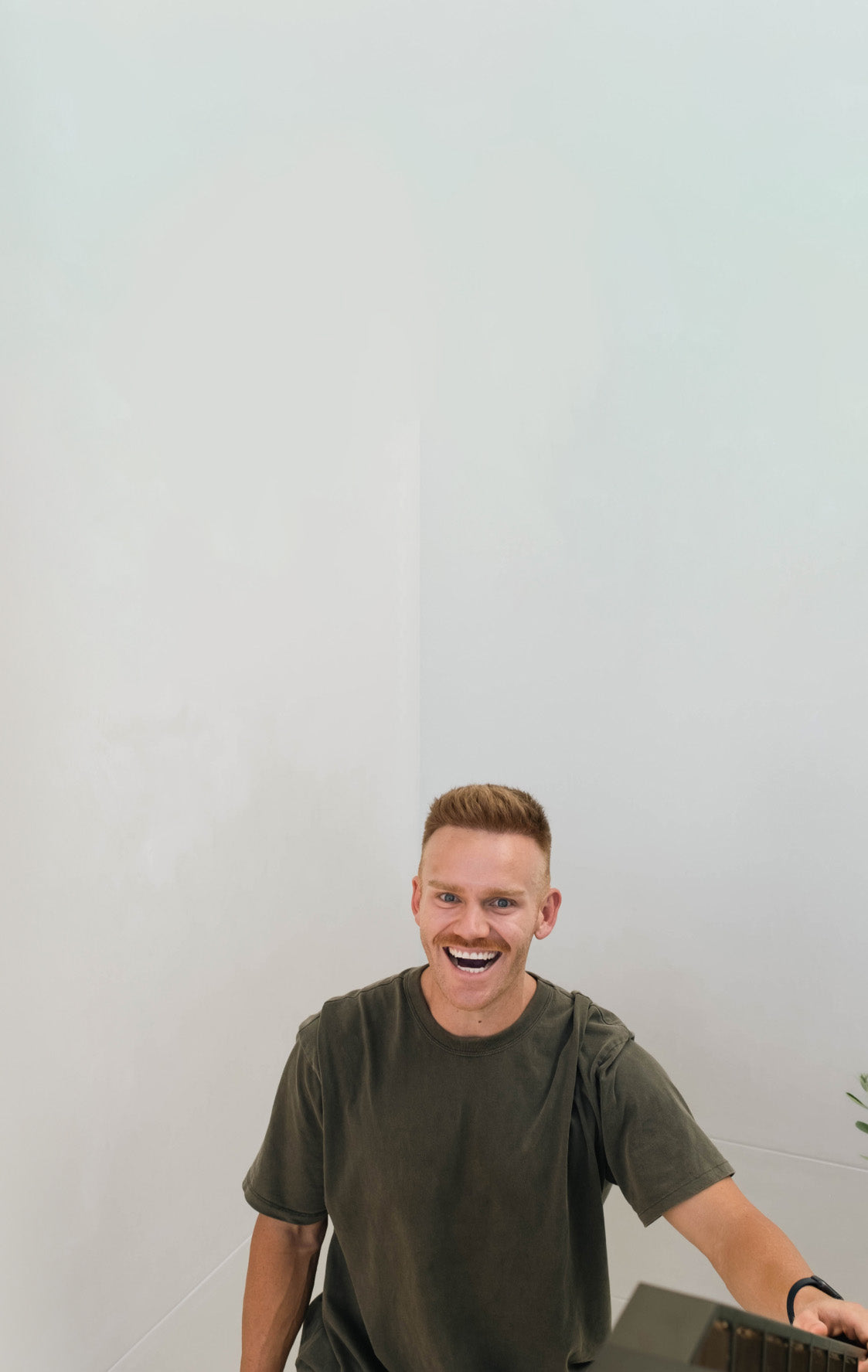 A person with short, light hair is smiling widely while sitting in a minimalist room with light-colored walls. They are wearing a dark green t-shirt and looking towards the camera. There is a small green plant partly visible on the right side.