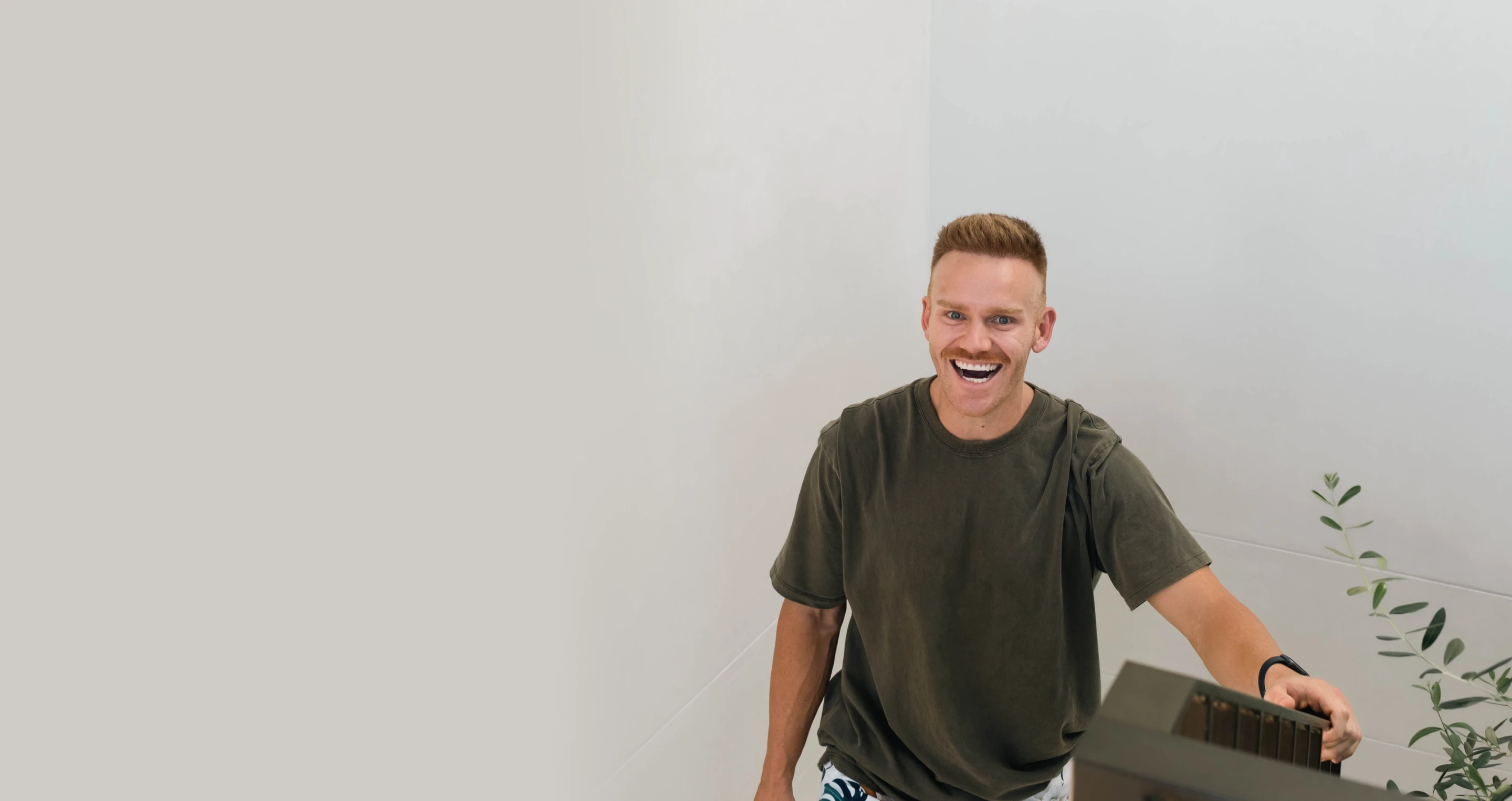 A smiling man with short hair and a mustache, wearing a dark t-shirt and patterned shorts, looking up while seated in a modern, minimalist indoor space. A potted plant is partially visible on the right side, and the background is a light gray.
