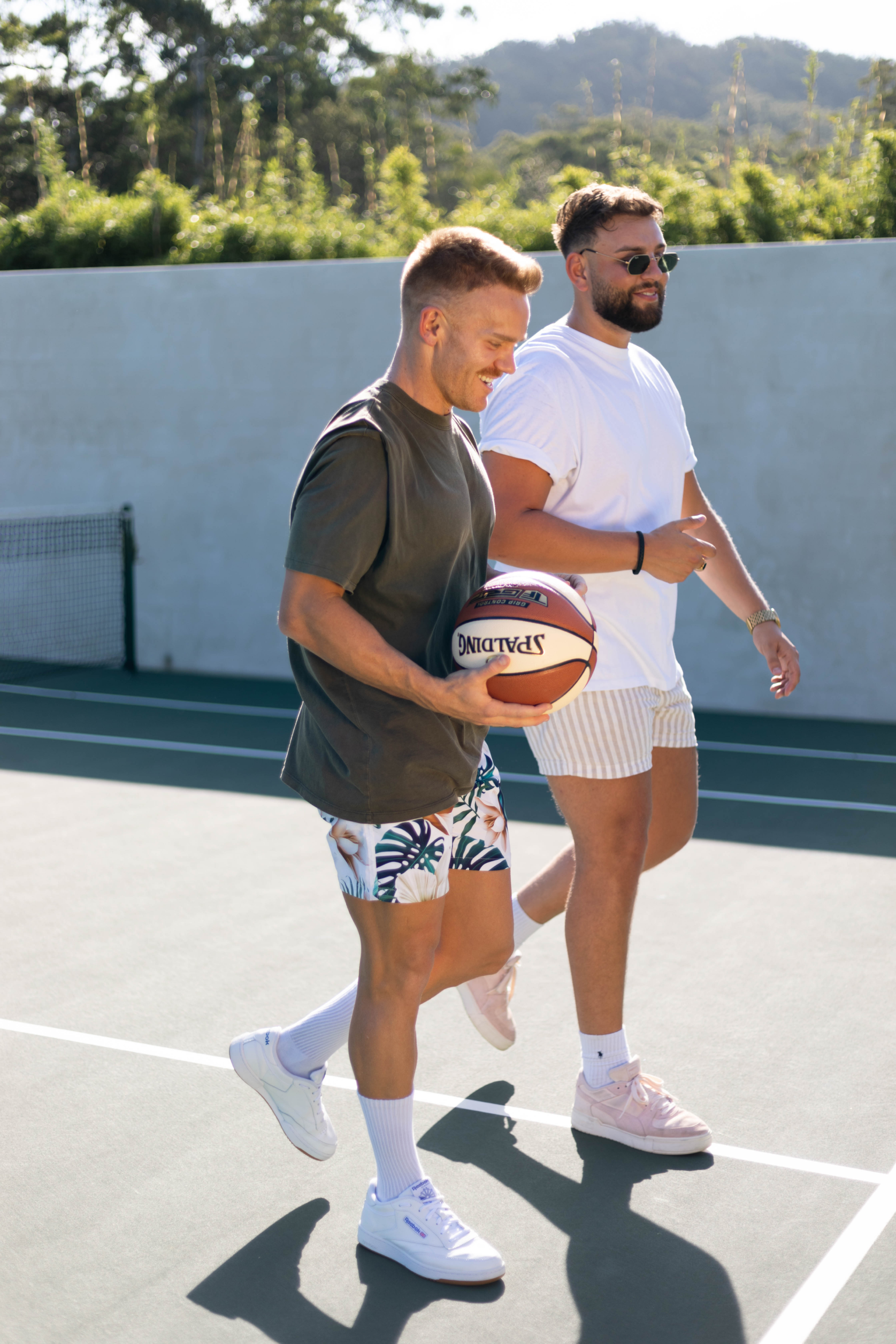 Two men walk side by side on an outdoor basketball court. One is holding a basketball, wearing a grey t-shirt and patterned shorts. The other wears a white t-shirt and light shorts. Both appear relaxed and are in casual summer attire, with a scenic backdrop of greenery.