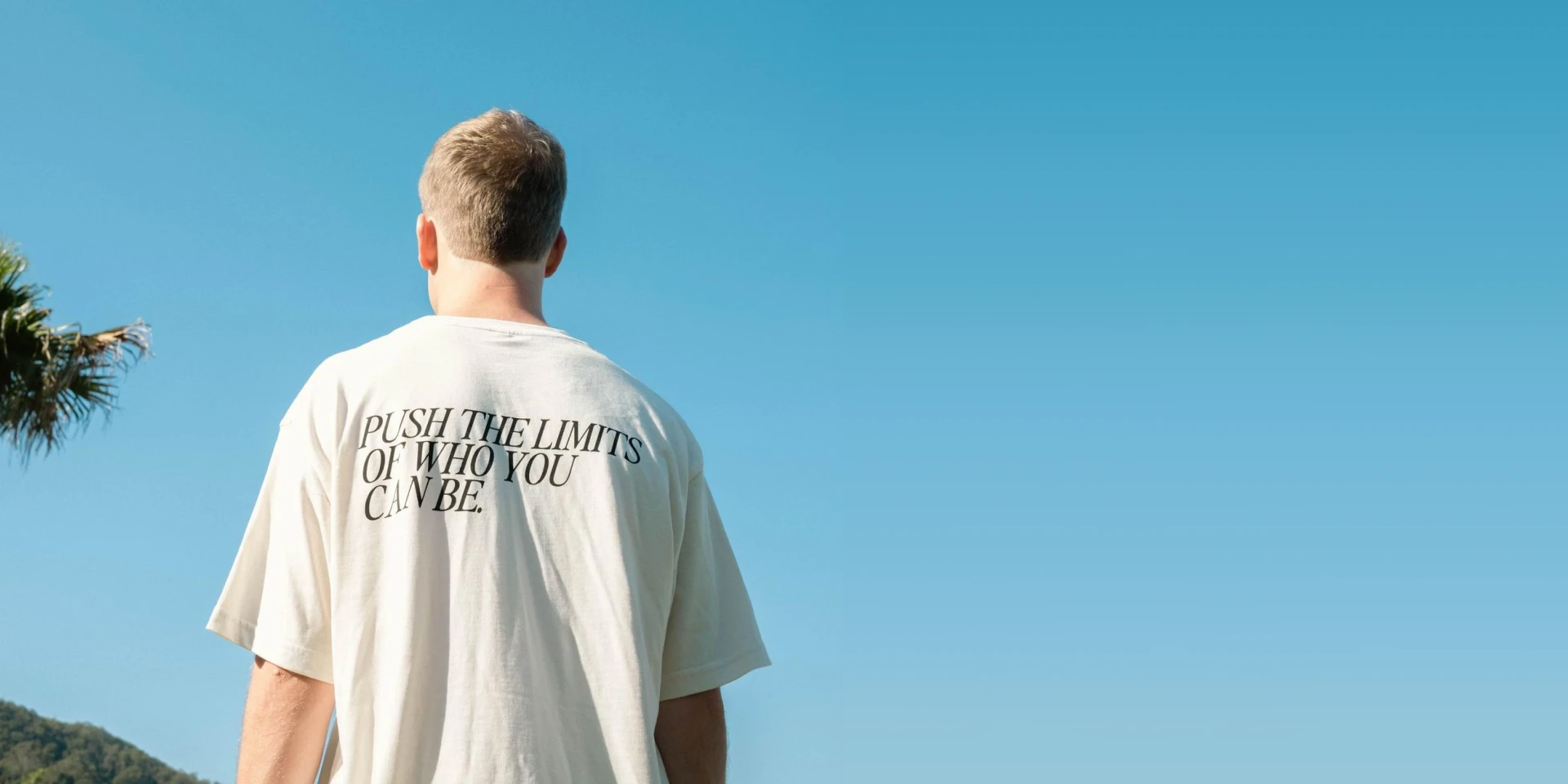 A person with short hair is standing with their back to the camera, wearing a white T-shirt that reads "Push the limits of who you can be." In the background, there is a clear blue sky and part of a tree and a hillside.